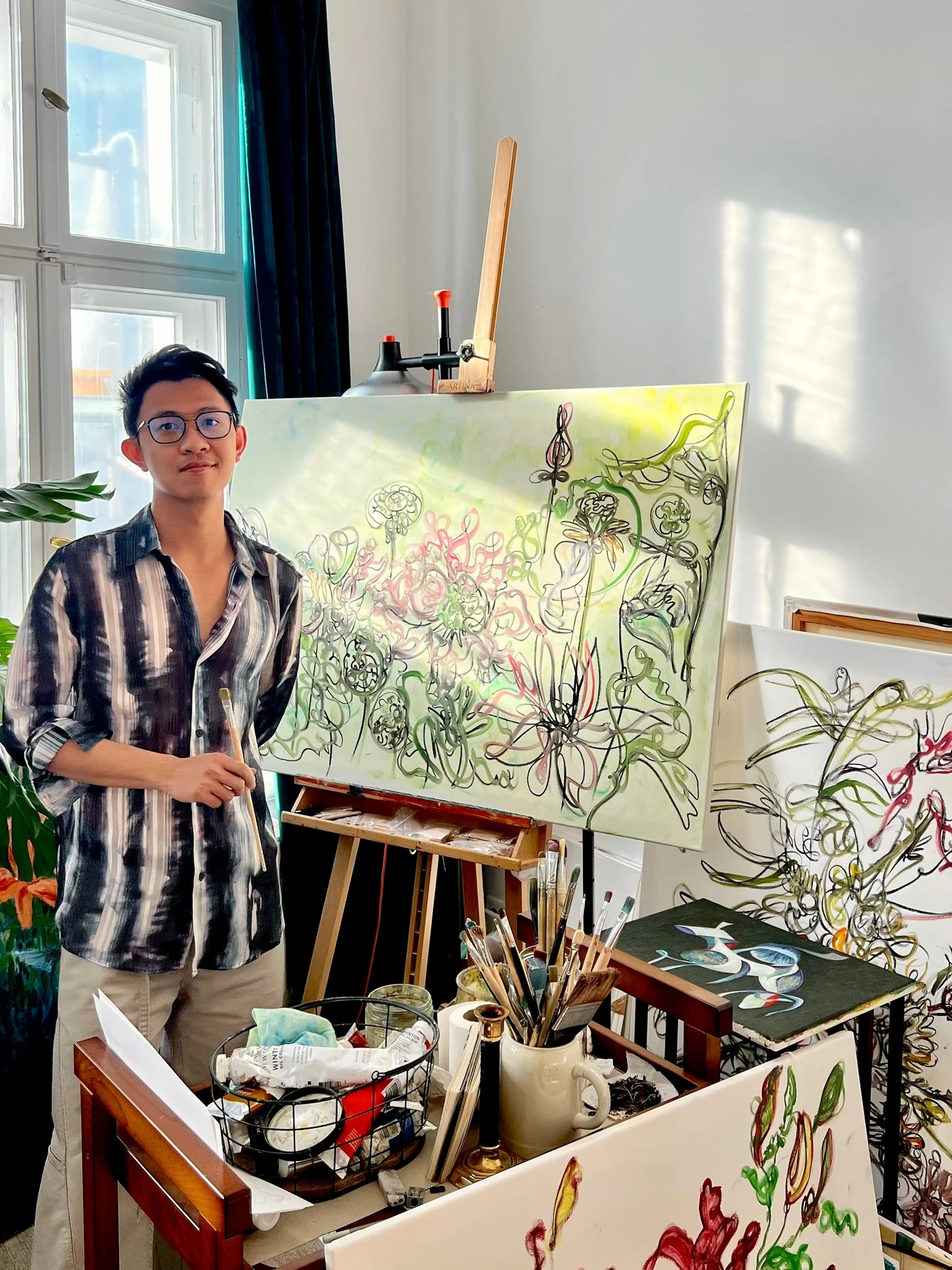 Dan Khoa standing in his studio beside floral paintings, an easel and painting materials.